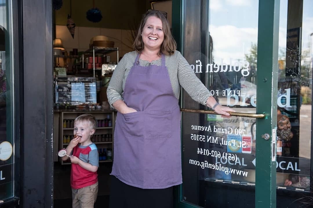 A woman wearing an apron stands in a doorway as a child eats with a spoon near the entrance to a bakery, blurred garden and storefront.