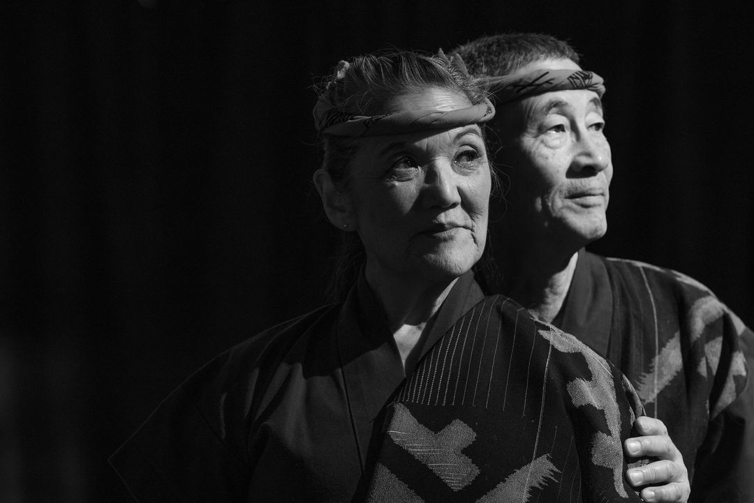 Monochrome portrait of an elderly Asian couple in traditional clothing, standing close against a dark backdrop, capturing cultural heritage.