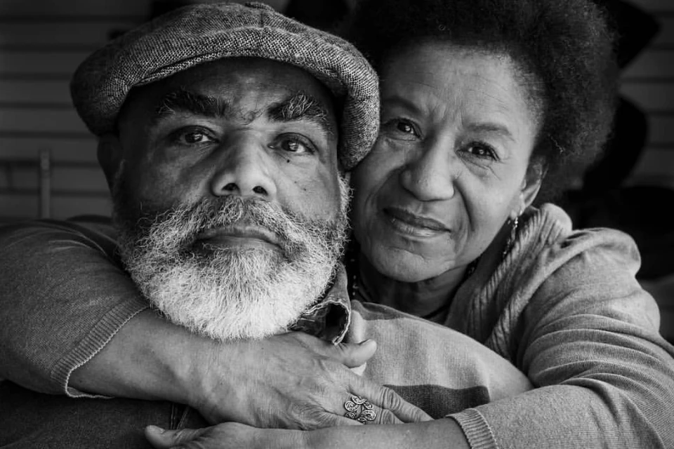 Monochrome portrait of an African-American couple in a close embrace, featuring an adult male in a hat and an adult female with curly hair.