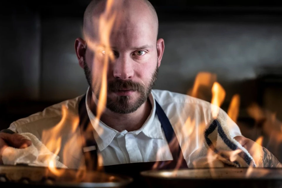 Chef with beard prepares food with flames rising in kitchen. Man wears white shirt and dark apron, cooking in an indoor environment.