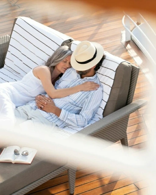 Mature couple lounging on a deck, embracing in the sun. She wears a white dress, he wears a blue shirt and hat, relaxing together outdoors.