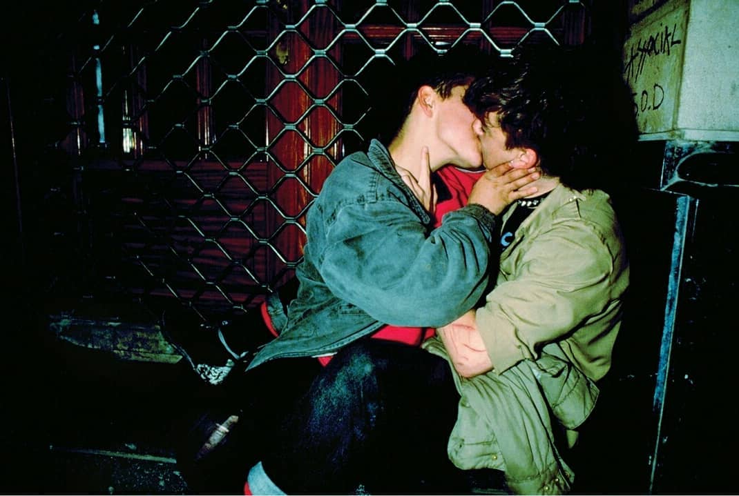 Two young men share a kiss in an urban environment at night, dressed in casual streetwear with a dark gate visible behind them.