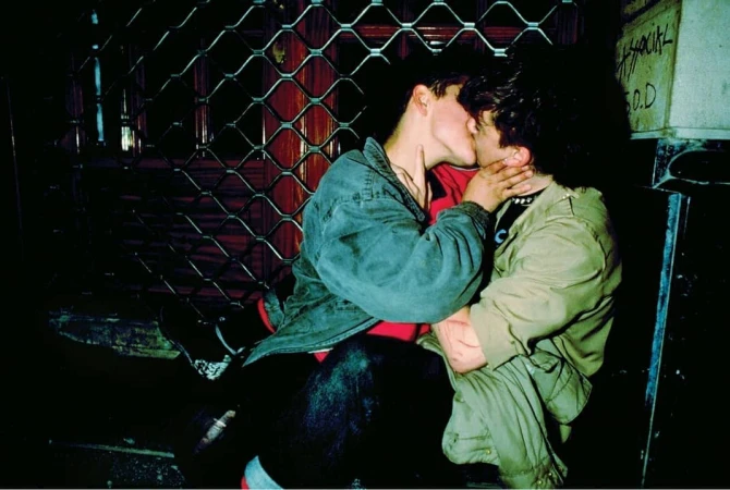 Two young men share a kiss in an urban environment at night, dressed in casual streetwear with a dark gate visible behind them.
