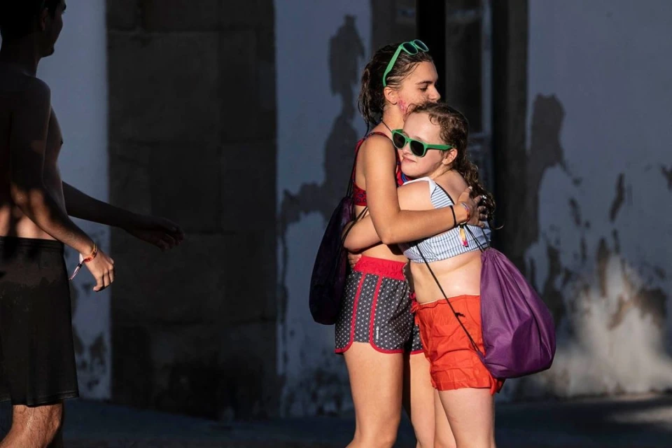Two young girls embrace outdoors on a sunny day, wearing sunglasses and swimsuits, carrying tote bags, and standing near a textured wall.