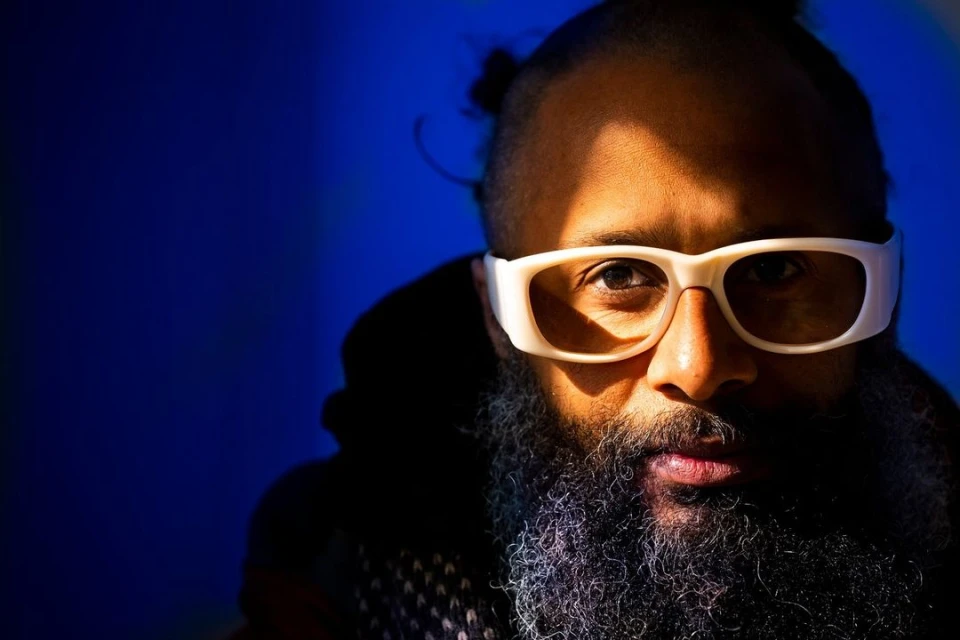 Close-up portrait of an adult with a salt-and-pepper beard, wearing white-rimmed glasses against a dark blue background with strong lighting.
