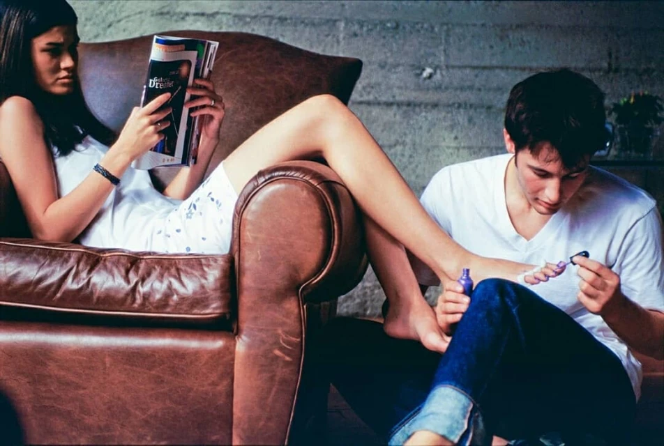Young man painting toenails on a woman relaxing in a brown leather armchair, while reading a magazine, in a casual indoor setting.