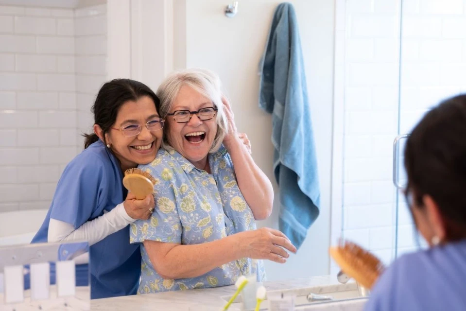 Elderly woman laughs as an aide hugs her and brushes her hair in the bathroom, reflecting in the mirror, fostering a heartwarming scene.