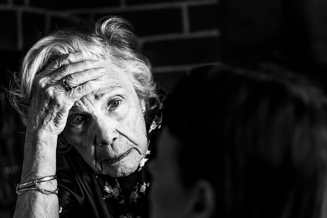 Monochrome portrait of an elderly woman with her hand on her forehead, facing another person in a dimly lit indoor setting.
