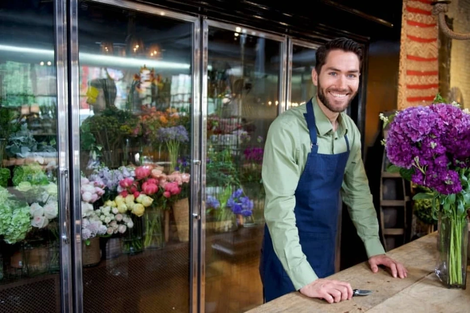 A smiling florist stands in front of a floral display cooler, wearing an apron, with a vase of purple hydrangeas on the wooden counter.