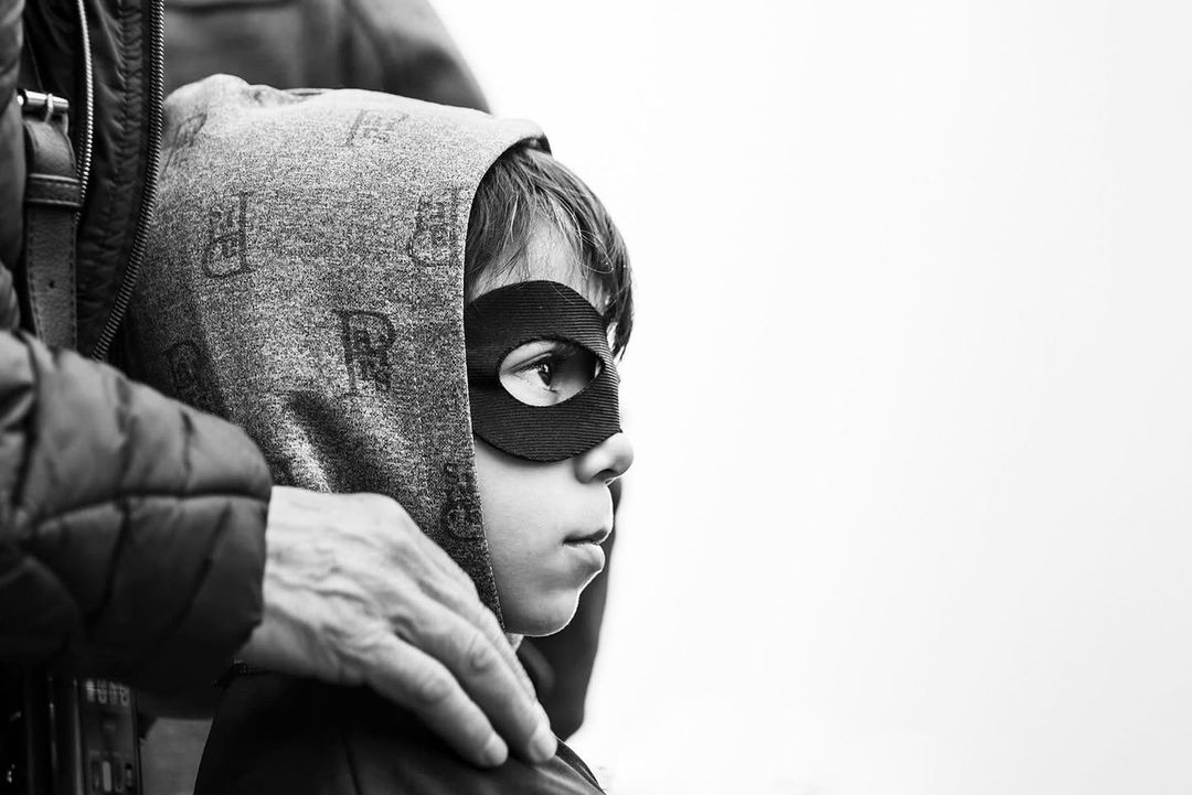 Close-up black and white portrait of a child in a superhero mask and hooded jacket with an adult's hands on their shoulders.