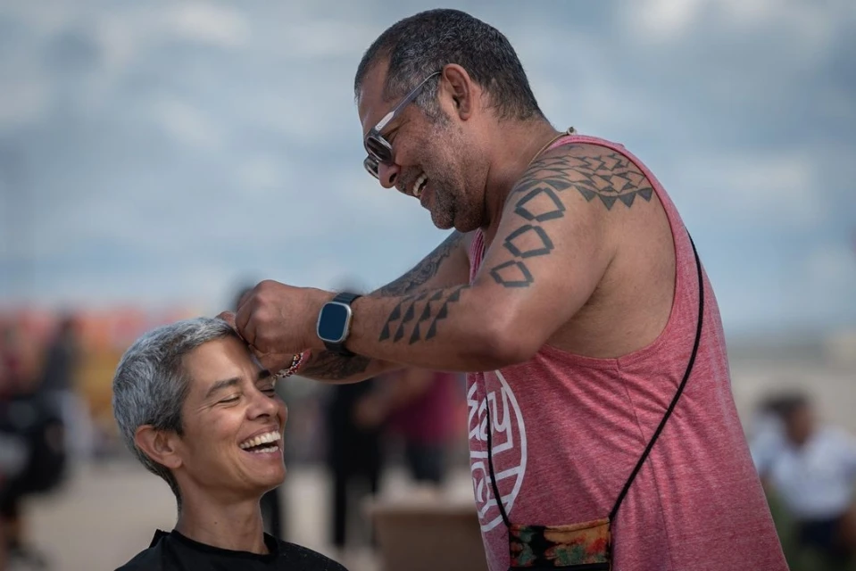 Two adults outdoors: a man gives a woman a haircut; both are smiling against a blurred background of beach and sky on a sunny day.