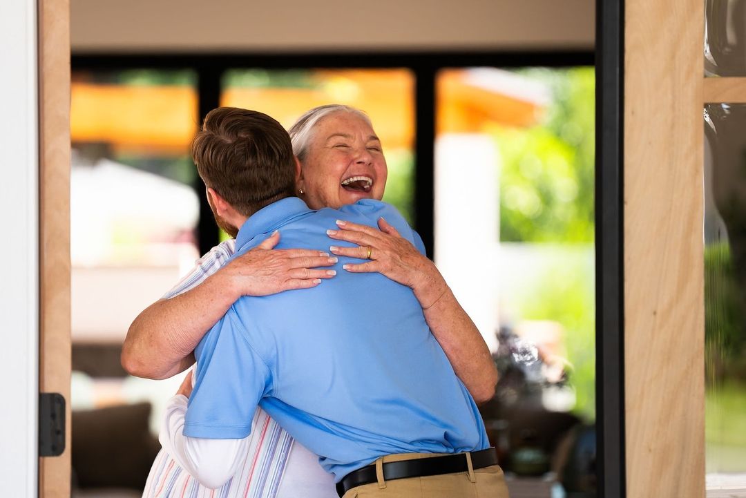 An adult woman hugs a younger adult man indoors near a doorway with natural light and greenery visible through the door's window.