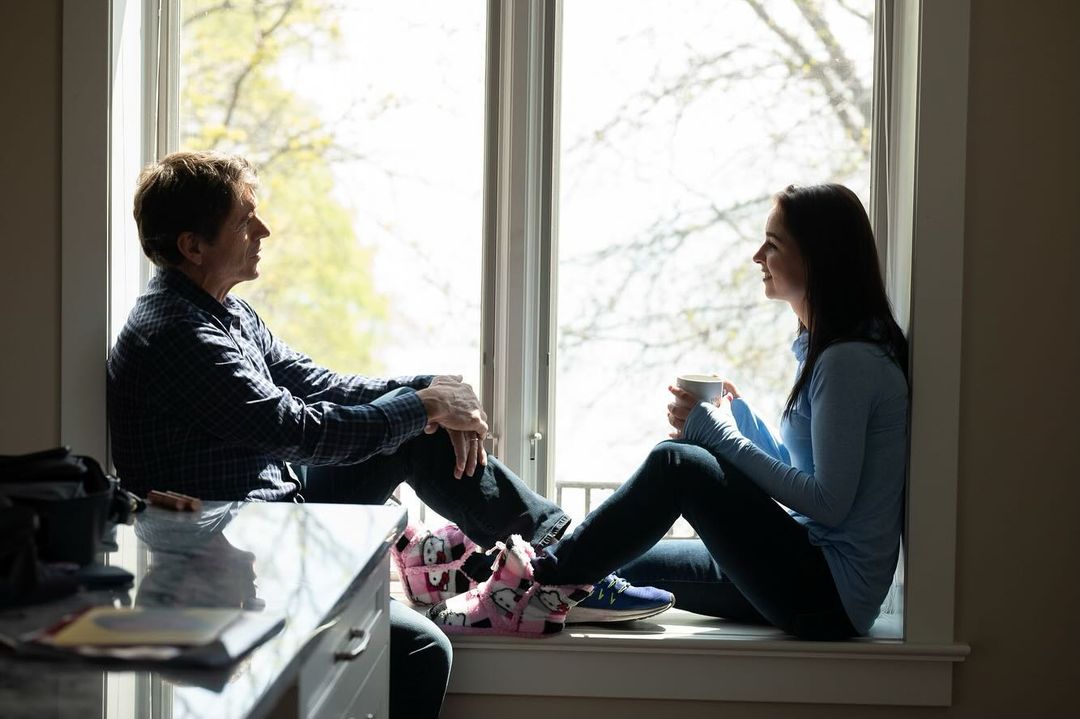 Man and woman sit in window of home interior in casual clothing, bright daylight coming through window. The pair are conversing casually.