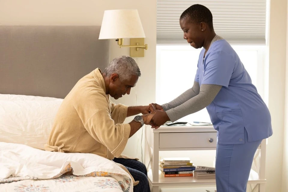 An elderly man in a bathrobe is assisted by a healthcare worker wearing scrubs in a bedroom setting near a bedside table.