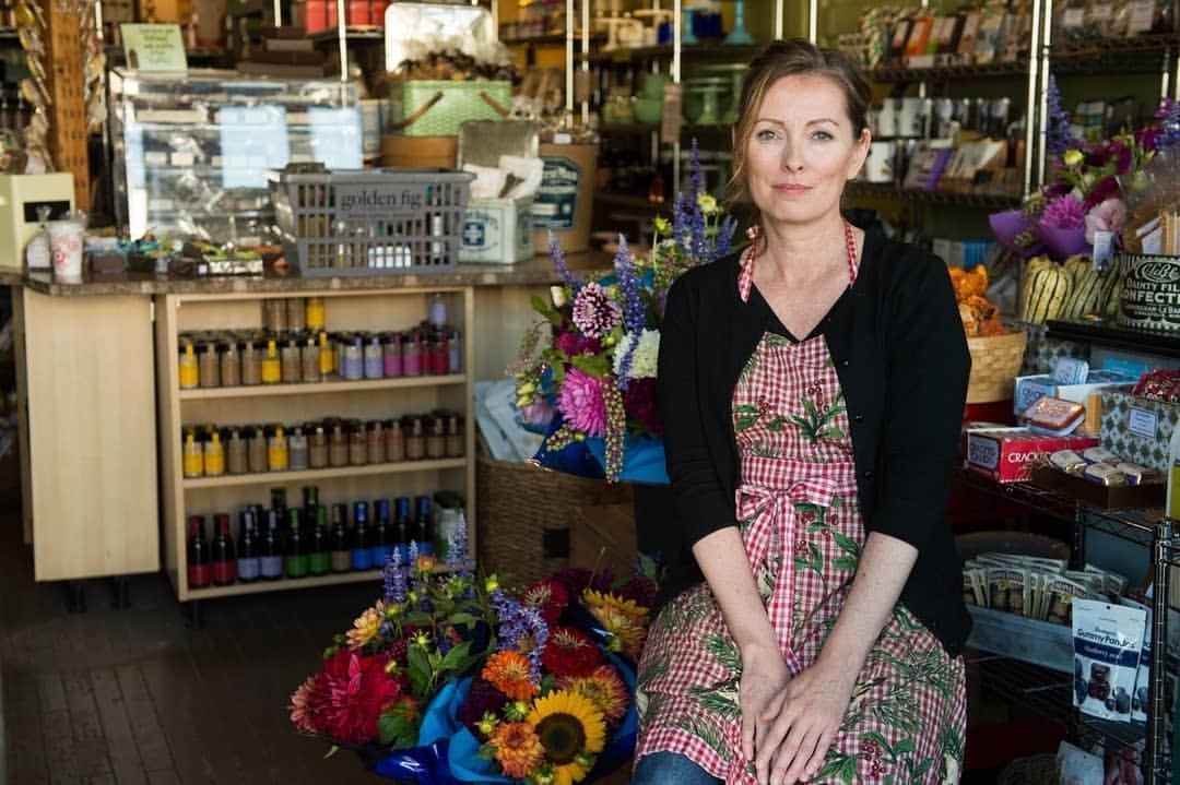 Woman in a red and white checkered apron sitting in a shop surrounded by flowers and goods on shelves, with natural window lighting.