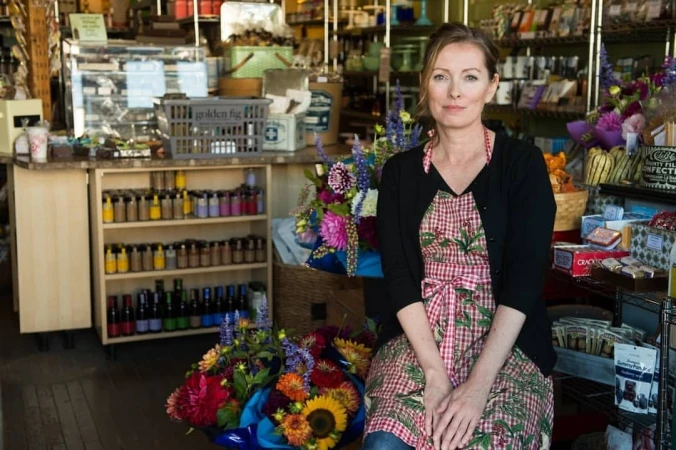 Woman in a red and white checkered apron sitting in a shop surrounded by flowers and goods on shelves, with natural window lighting.