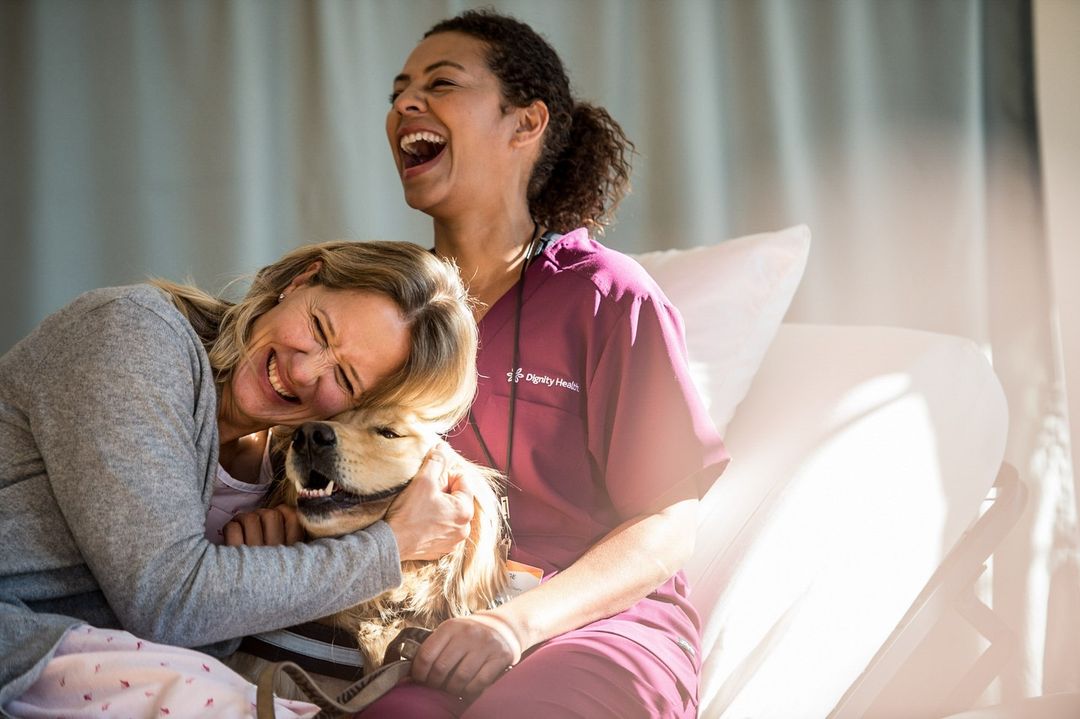 Woman hugs a golden retriever with a healthcare worker joyfully laughing in the background, creating a heartwarming scene in the hospital room.