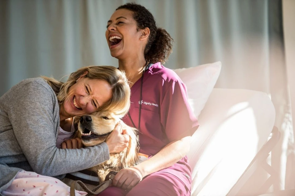Woman hugs a golden retriever with a healthcare worker joyfully laughing in the background, creating a heartwarming scene in the hospital room.