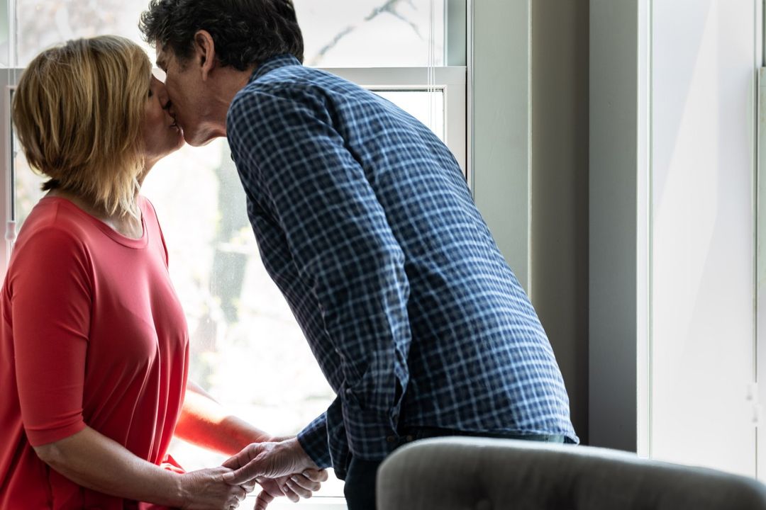 A couple shares a kiss in front of a window, holding hands and creating a tender moment of connection and affection in a bright indoor space.