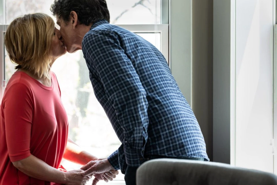 A couple shares a kiss in front of a window, holding hands and creating a tender moment of connection and affection in a bright indoor space.