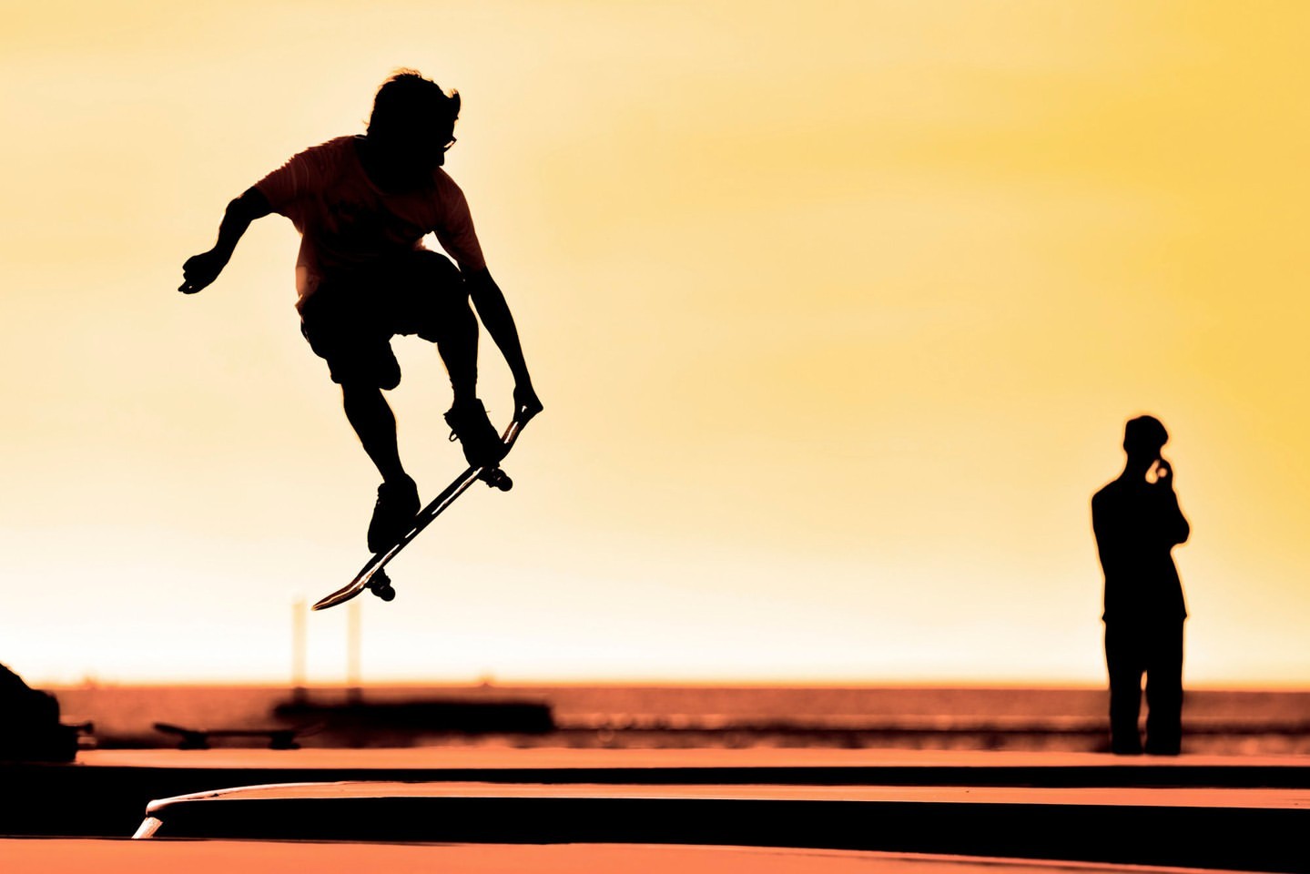 Silhouette of a skateboarder performing a jump trick against an orange sky at dusk, with a second person watching in the distance.