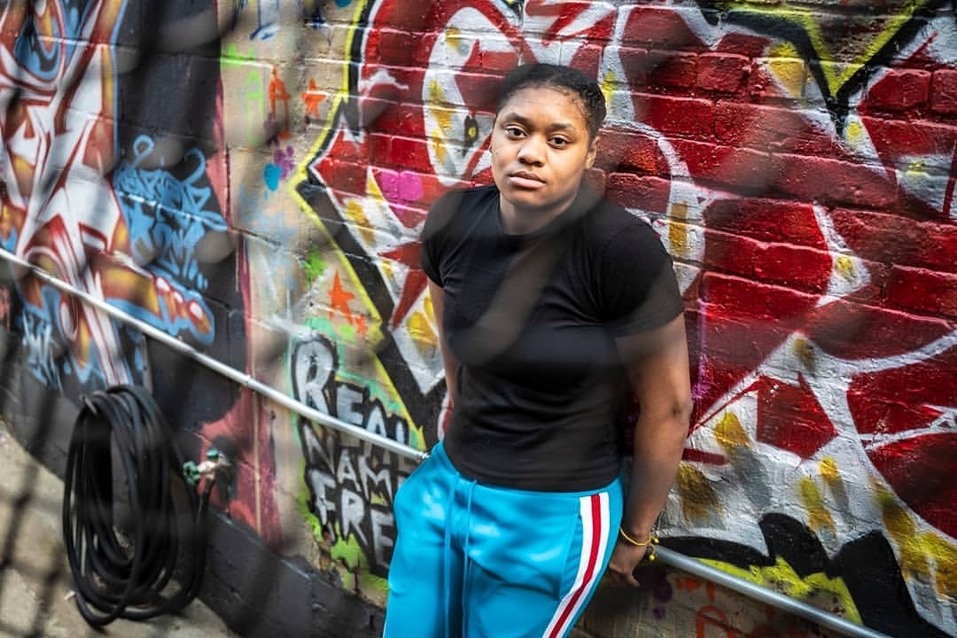 Young woman poses in black top and blue track pants against graffiti wall, captured in a medium shot outdoors during daylight.