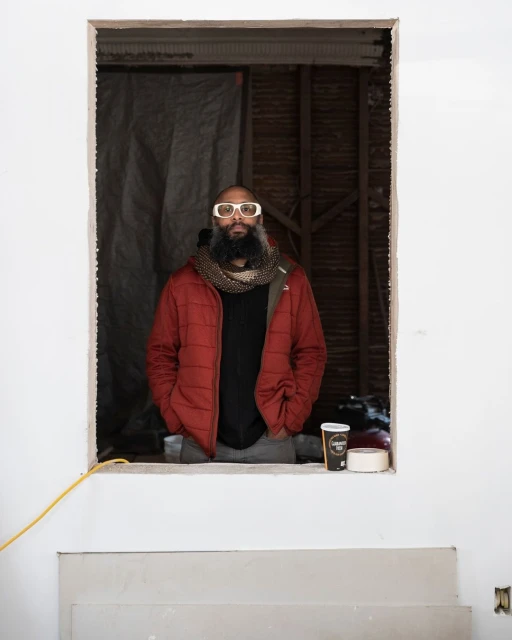 Adult male with a beard wearing sunglasses and a red puffer jacket standing inside a room with unfinished walls and exposed wood framing.