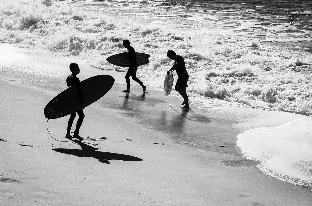 Three surfers carrying surfboards walk along a sandy beach near crashing ocean waves in a monochrome photograph.
