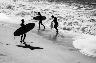 Three surfers carrying surfboards walk along a sandy beach near crashing ocean waves in a monochrome photograph.
