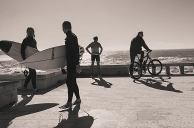 Four men near a coastal promenade with two surfers carrying surfboards, a standing figure, and a cyclist by the ocean.
