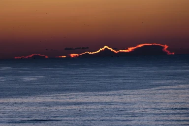 Vibrant fiery sunset cloud formation glowing red above dark ocean water with distant horizon and birds in the sky.