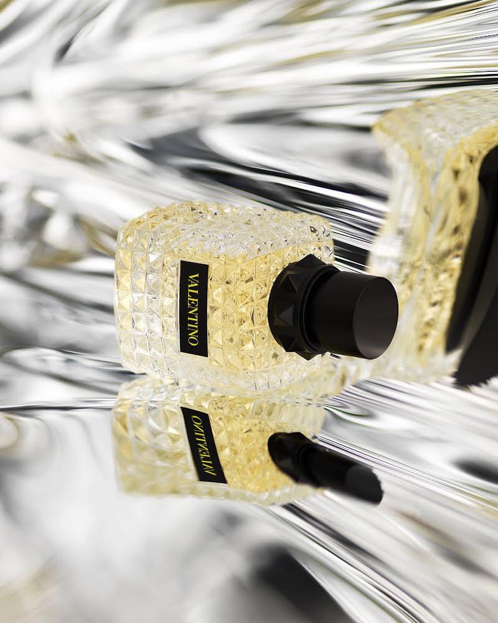 Valentino perfume bottle displayed on a reflective surface, crystal clear bottle with black cap, elegant and luxurious fragrance.