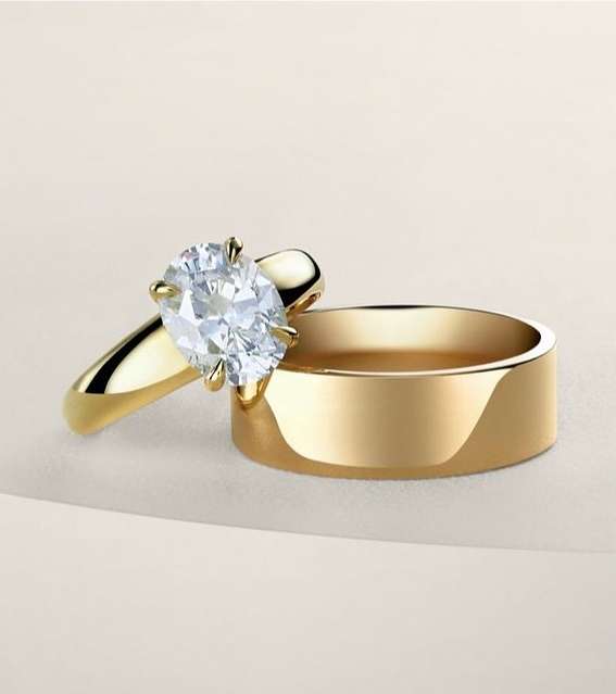 Close-up of two gold wedding rings, one with a large oval diamond, arranged on a soft gradient beige background with studio lighting.
