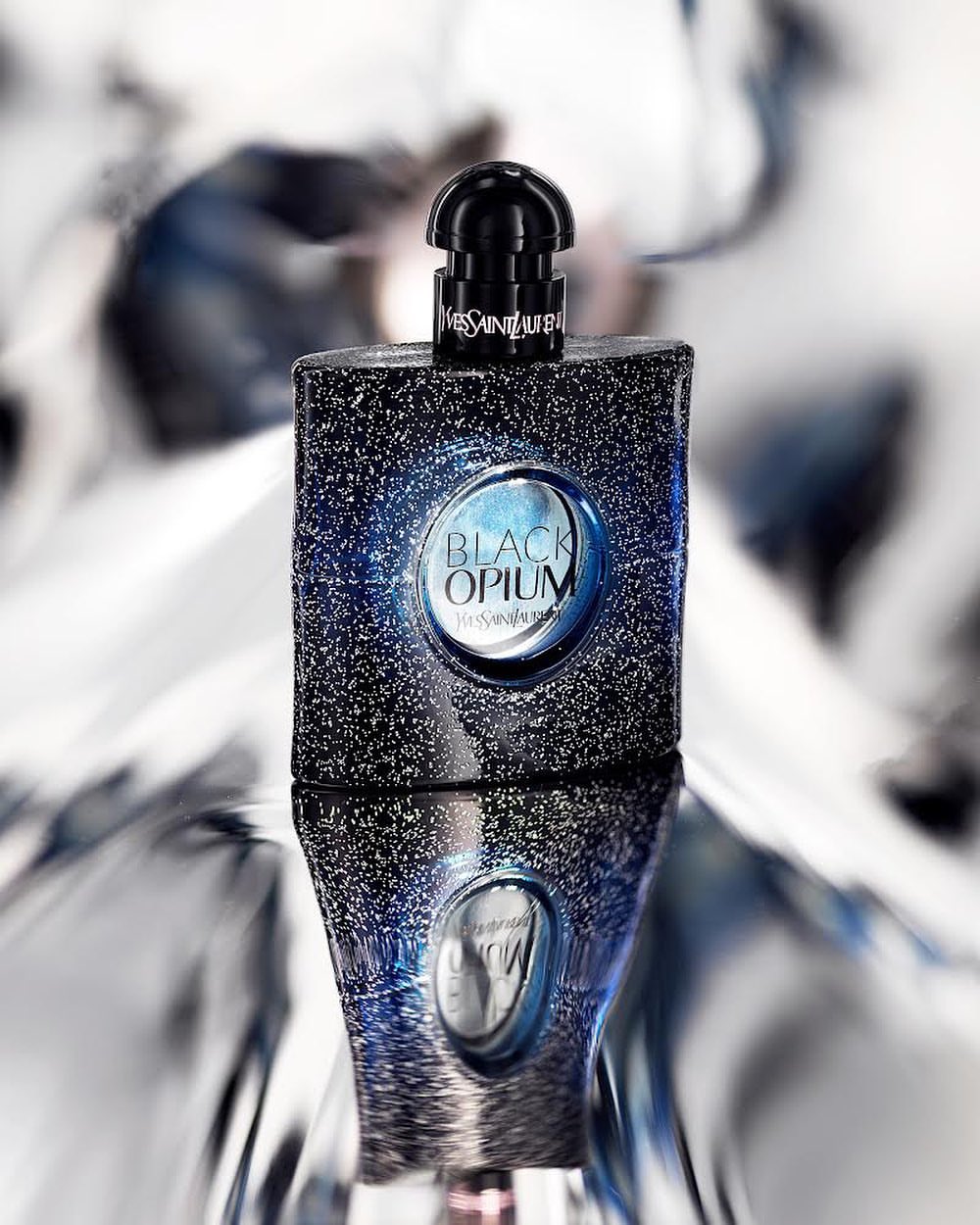 Black Opium perfume bottle by Yves Saint Laurent reflected against a shiny background. Bottle is glittery black with a round label.