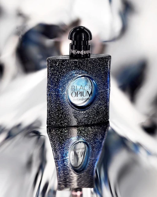 Black Opium perfume bottle by Yves Saint Laurent reflected against a shiny background. Bottle is glittery black with a round label.