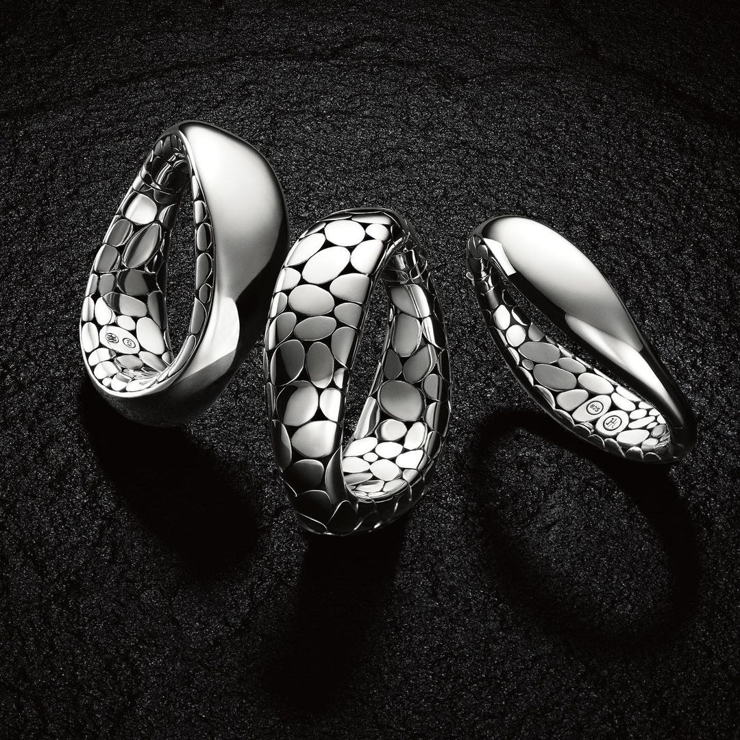 Three silver-colored bangles with oval-shaped inner designs are arranged on a black textured surface, reflecting light and shadows.