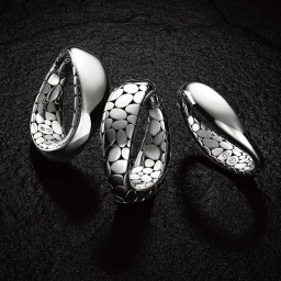 Three polished silver hoop earrings with textured pebble pattern interiors arranged on a dark rough stone background