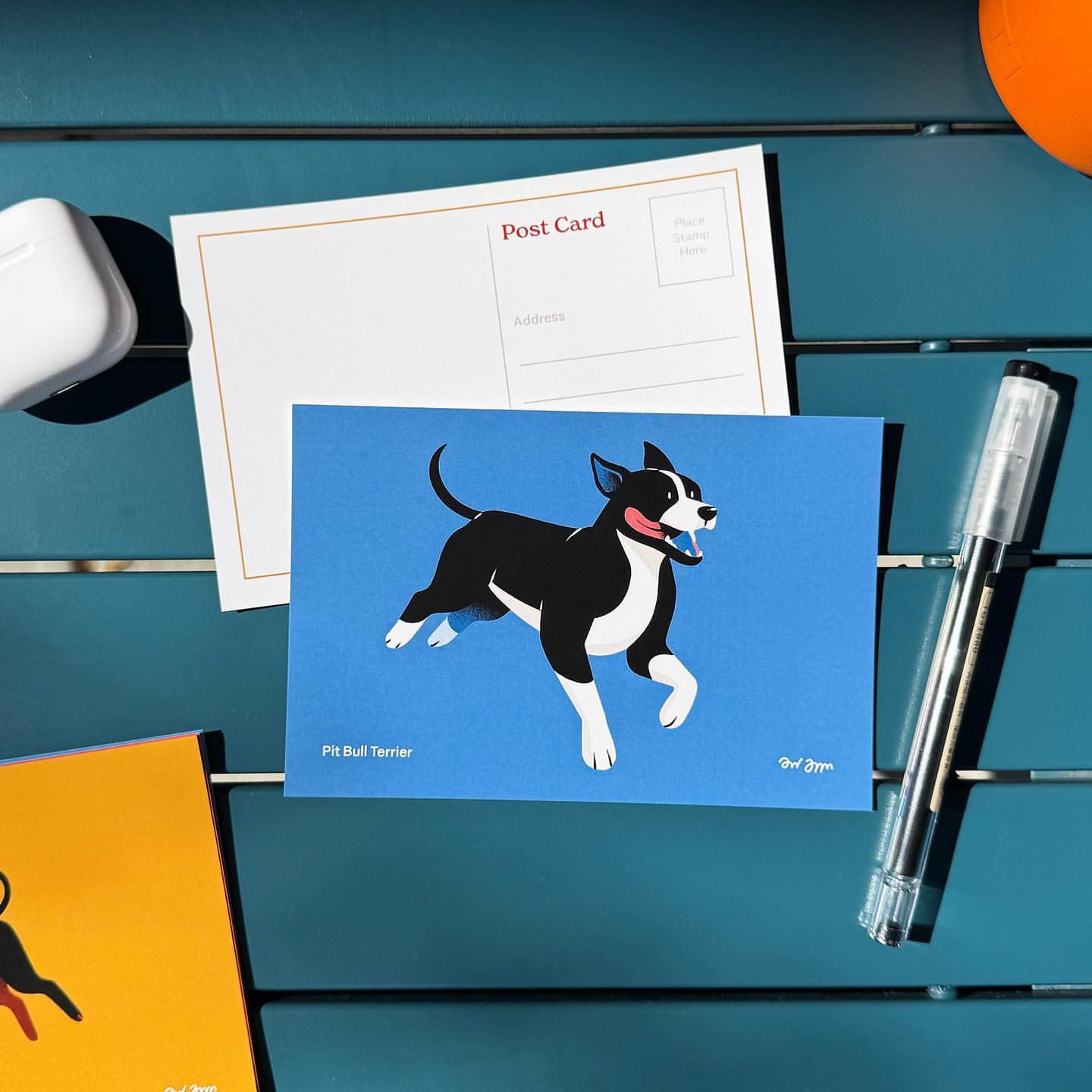 Illustration of a black and white Pit Bull Terrier dog on a blue card, displayed with a postcard, pen, and case on a blue surface.