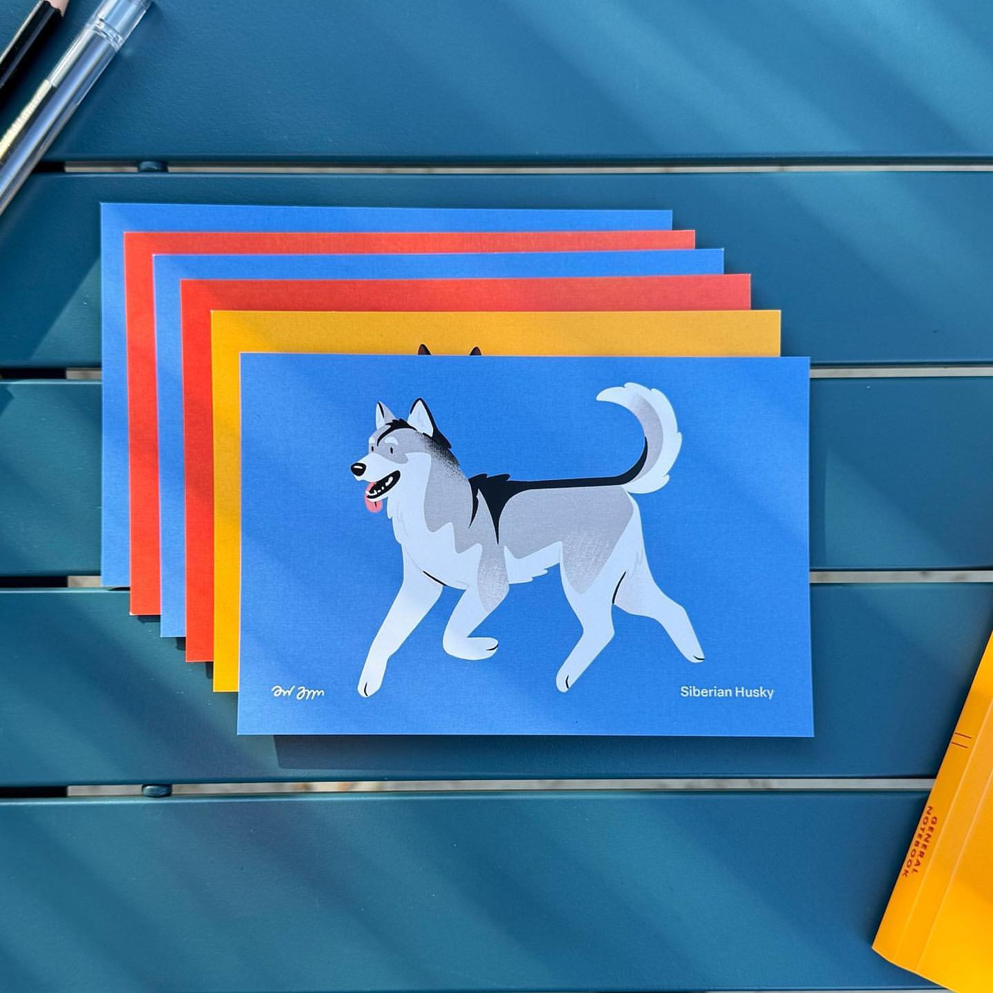 Illustration of a Siberian husky dog on a blue postcard, part of a stack of colorful postcards on a blue slatted surface.
