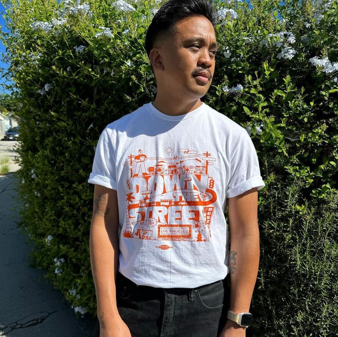 An adult Asian man wears a graphic T-shirt and poses outdoors near a green hedge in the bright sunlight, showcasing a casual style.