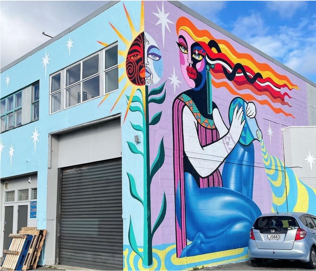 Vibrant mural art on a building's exterior featuring human figures, plants, celestial shapes, and bold colors in an urban setting.