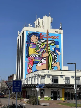 Mural art on an urban building featuring Maori-inspired figures and designs, set against a clear blue sky in an outdoor setting.