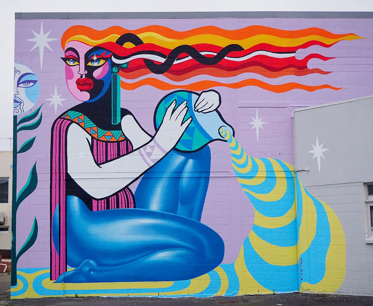 Vibrant mural art on an exterior wall featuring a stylized woman pouring liquid from a patterned vessel, set against a light purple backdrop.