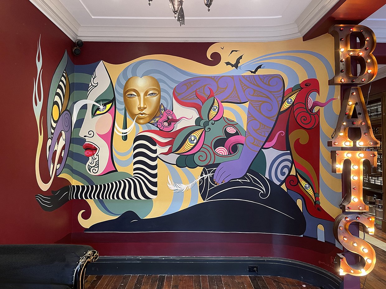 Eye-level shot of a vibrant mural featuring abstract figures, golden mask, and bold colors, complemented by illuminated BATS sign.