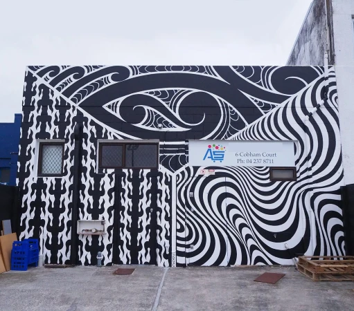 Building exterior showcasing Maori-inspired mural with black and white patterns, including spiral designs and geometric elements under a cloudy sky.