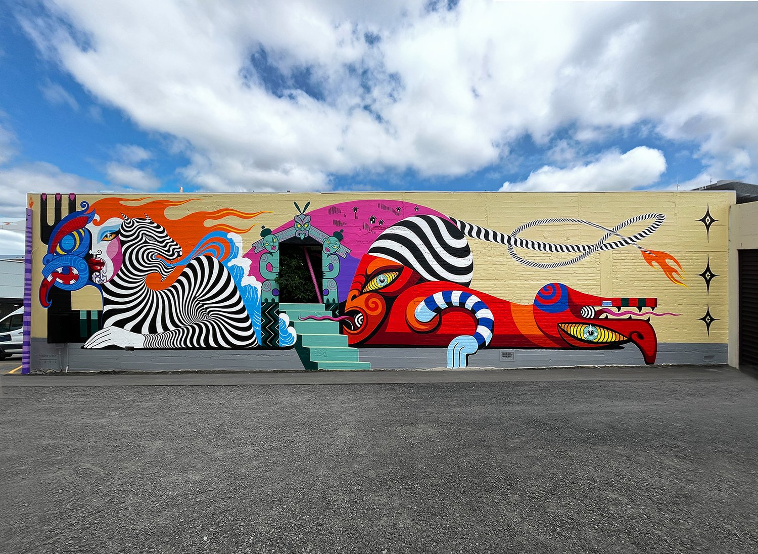 Eye-level shot of a colorful mural on a building with fantastical figures, stairs, and a blue sky with white clouds in the background.