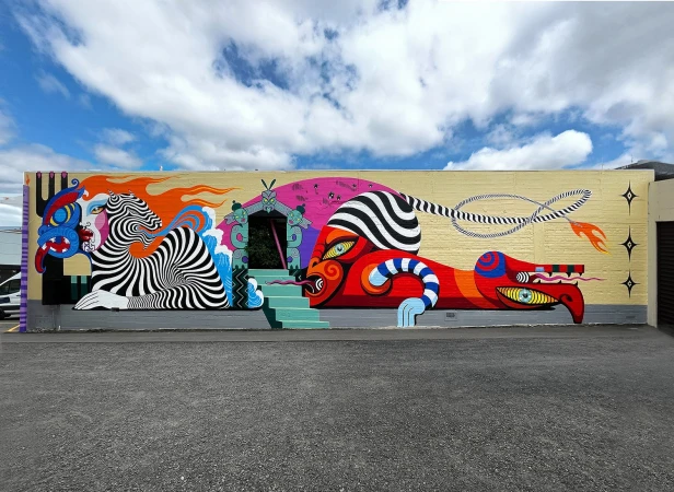 Eye-level shot of a colorful mural on a building with fantastical figures, stairs, and a blue sky with white clouds in the background.