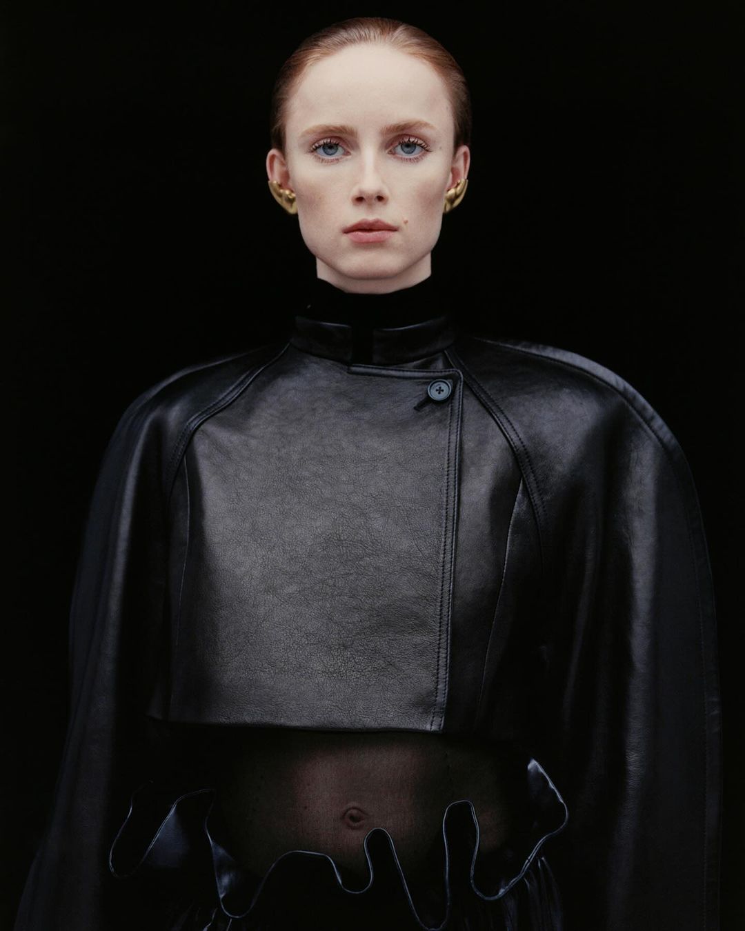 Woman in a black leather capelet and top with unique hem detail poses against a dark background, showcasing minimal makeup and gold earrings.