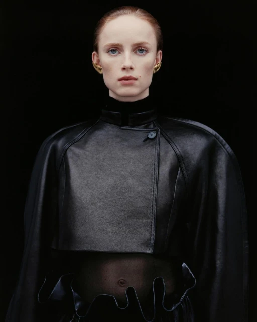Woman in a black leather capelet and top with unique hem detail poses against a dark background, showcasing minimal makeup and gold earrings.