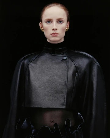 Woman in a black leather capelet and top with unique hem detail poses against a dark background, showcasing minimal makeup and gold earrings.